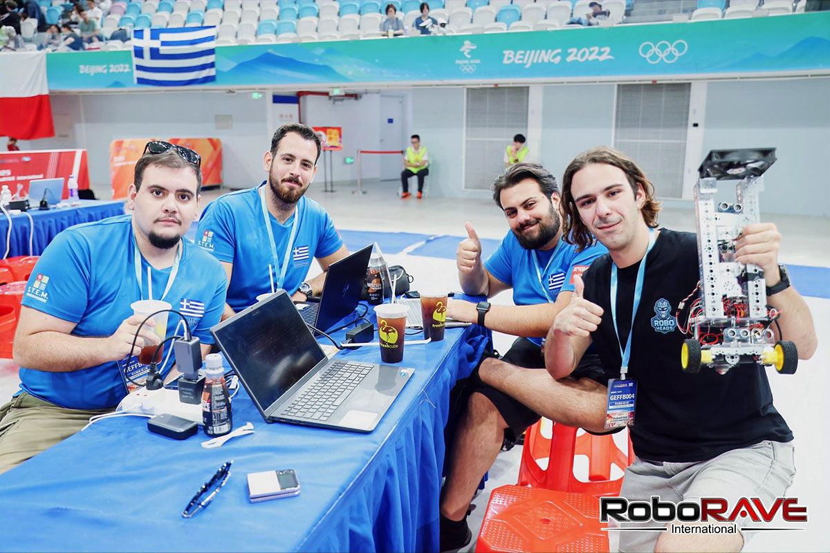 Η ομάδα στο τραπέζι εργασίας της με ανοιχτούς φορητούς υπολογιστές και το πυροσβεστικό ρομπότ, κάνοντας το σήμα της επιδοκιμασίας, με μια ελληνική σημαία κρεμασμένη στον τοίχο πίσω τους.