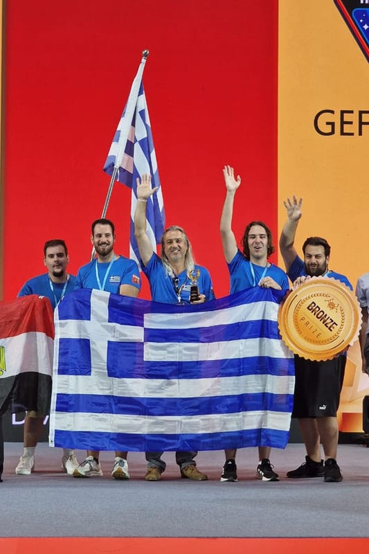 Η ομάδα στη σκηνή με μετάλλια και την ελληνική σημαία ανοιχτή, πανηγυρίζοντας μπροστά σε πανό που δείχνει ένα μεγάλο χάλκινο μετάλλιο.