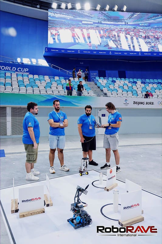 Τέσσερα μέλη της ομάδας Roboheads με μπλε μπλούζες στέκονται δίπλα στο ρομπότ τους πάνω στη λευκή αγωνιστική πίστα, με τις κερκίδες και μια μεγάλη οθόνη πίσω τους.