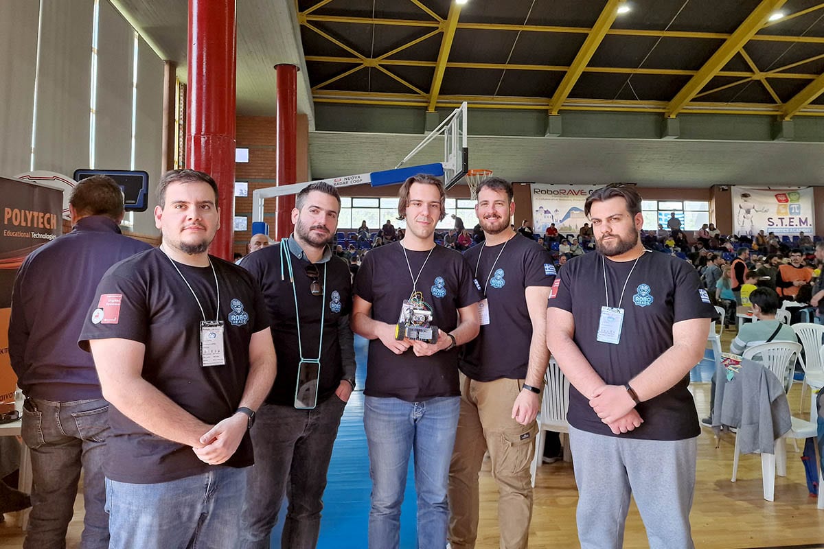 Η ομάδα Roboheads στέκεται στην αίθουσα του διαγωνισμού, με ένα μέλος να κρατά το μικρό αυτόνομο ρομπότ λαβυρίνθου και το πανό του RoboRAVE Greece πίσω τους.